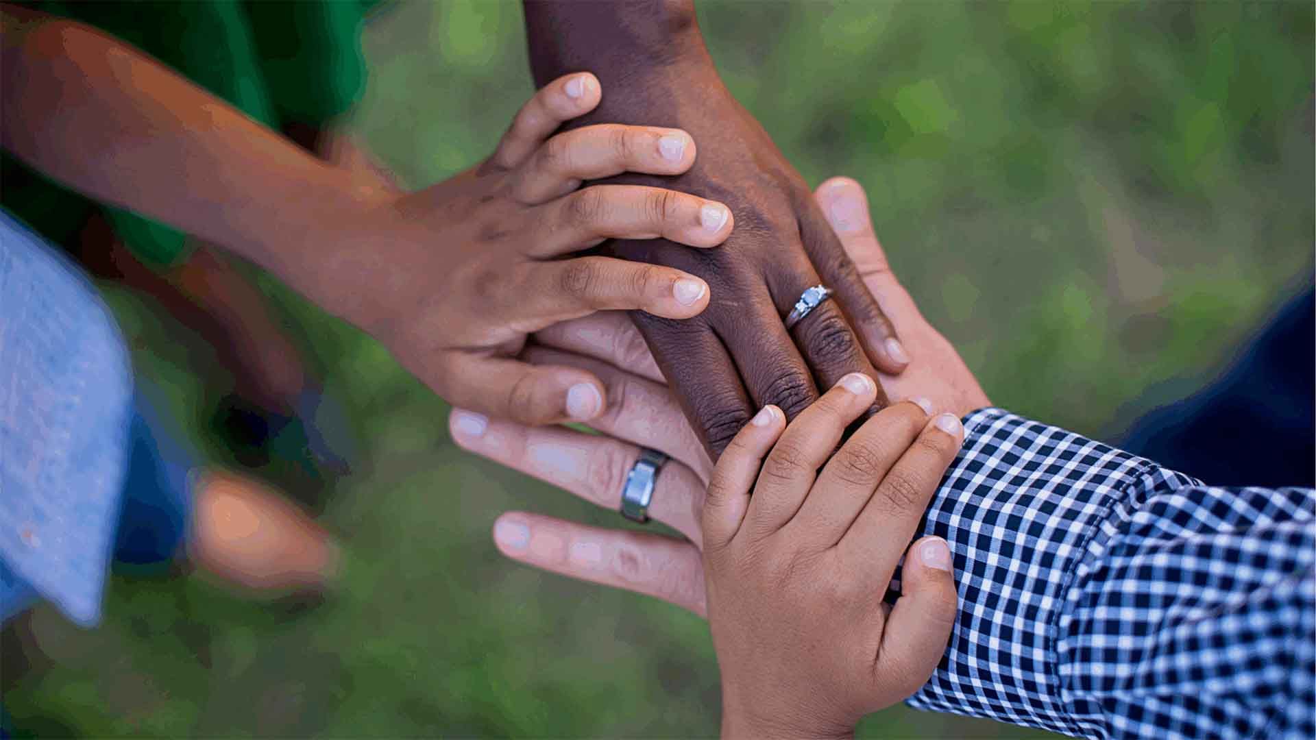 An overhead, close-up shot of the hands of a multi-racial family stacked together in a gesture of unity. The hands of a Black person and a white person, both wearing wedding rings, are layered with the smaller hands of their children over a soft green background.