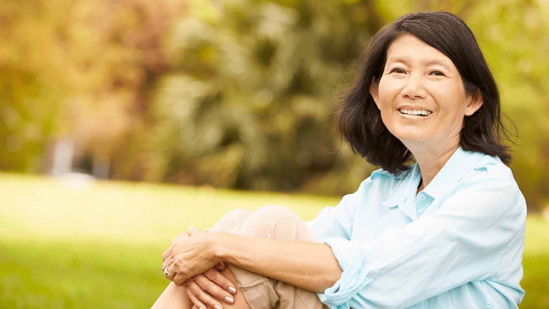 A happy, middle-aged East Asian woman with short dark hair sits on a grassy lawn in a park. She is smiling warmly as she hugs her knees, looking relaxed and content.