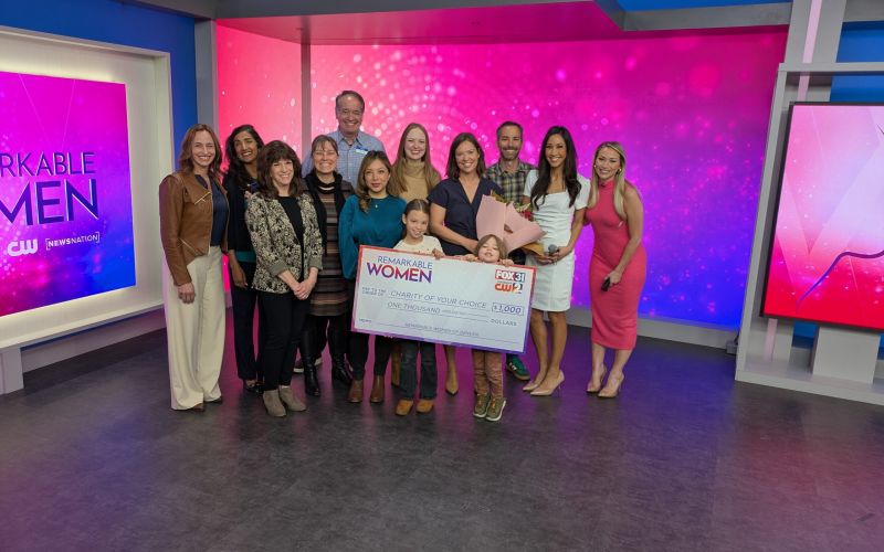 A large group of people poses for a photo on a television studio set for the "Remarkable Women" awards. In the center, a woman, surrounded by family, friends, and TV personalities, holds a large ceremonial check for $1,000 for a "Charity of Your Choice" from FOX31 and CW Channel 2.