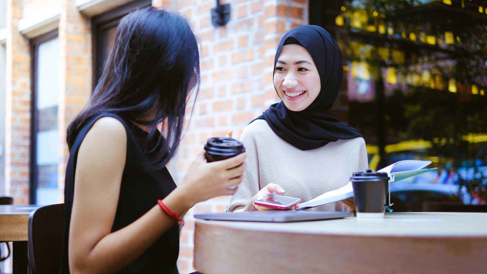 Two women are sitting at an outdoor cafe table, engaged in a friendly conversation. One woman, wearing a black hijab and a light-colored sweater, is smiling brightly at the other woman.